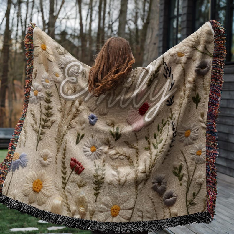 May include: Beige throw blanket with embroidered floral design and the name "Emily" in white script. The blanket features various flowers in white, yellow, red, and blue, with green leaves. The edges have a fringe in multiple colors.