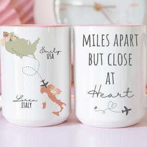 May include: White ceramic mug with a pink handle and rim. One side features a map of the USA and Italy with names and the text "Miles Apart But Close At Heart". The other side has the same text.