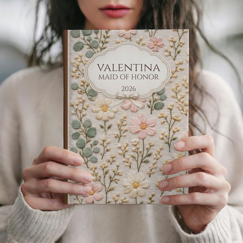 May include: A journal with a floral design and the text "VALENTINA MAID OF HONOR 2026". The cover features embroidered flowers in pink, cream, and green, with a cream-colored background. The journal has a brown spine.