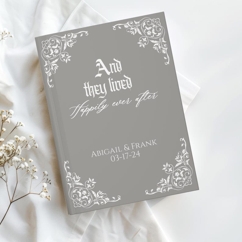 May include: A gray book with white floral corner designs and text that reads "And they lived Happily ever after." The names "Abigail & Frank" and the date "03-17-24" are also printed on the cover. The book is on a white surface.