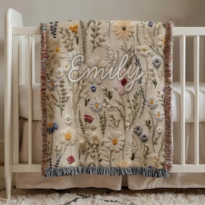 May include: A beige baby blanket with embroidered flowers in various colors, including white, yellow, blue, and red. The name "Emily" is embroidered in white script across the blanket. The blanket has a fringed edge and is draped over a white crib.