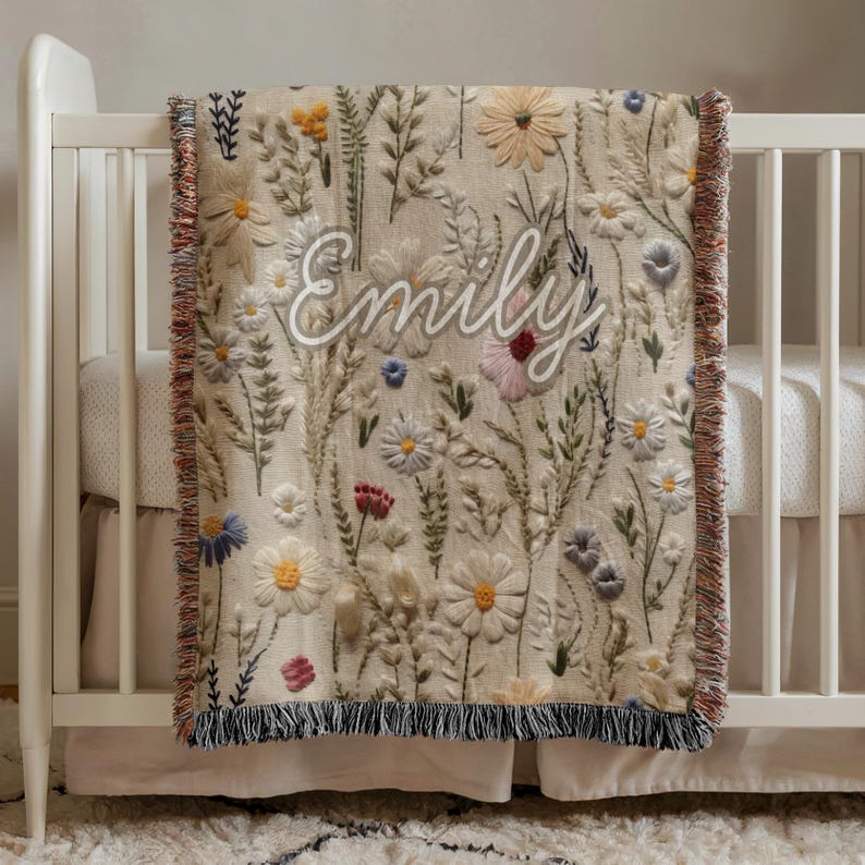 May include: A beige baby blanket with embroidered flowers in various colors, including white, yellow, blue, and red. The name "Emily" is embroidered in white script across the blanket. The blanket has a fringed edge and is draped over a white crib.