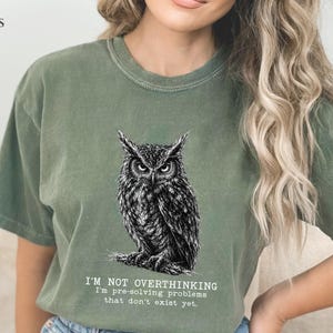 Puede incluir: Una camiseta de manga corta color verde musgo con una ilustraci&oacute;n detallada en blanco y negro de un b&uacute;ho posado en una rama. Debajo del b&uacute;ho, el texto dice: I'M NOT OVERTHINKING I'm pre-solving problems that don't exist yet. Cuello redondo.