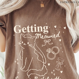 Puede incluir: Camiseta color espresso con el texto "Getting Meowied" y una ilustraci&oacute;n de gato. El dise&ntilde;o incluye anillos de boda, una copa de champ&aacute;n y corazones. Esta camiseta informal es perfecta para una despedida de soltera.