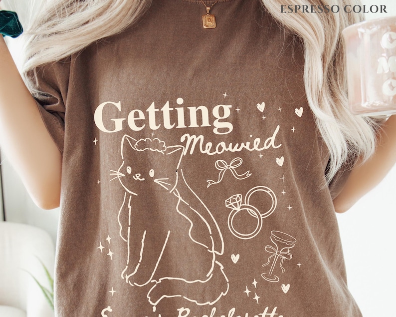 Puede incluir: Camiseta color espresso con el texto "Getting Meowied" y una ilustraci&oacute;n de gato. El dise&ntilde;o incluye anillos de boda, una copa de champ&aacute;n y corazones. Esta camiseta informal es perfecta para una despedida de soltera.