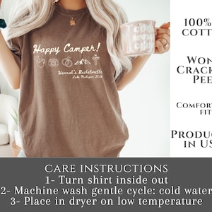 Puede incluir: Camiseta marr&oacute;n con el texto "Happy Camper!" e iconos de camping. El texto "Hannah's Bachelorette Lake Michigan 2026" est&aacute; debajo. Hecha de 100% algod&oacute;n estadounidense y producida en EE. UU. Tambi&eacute;n se incluyen instrucciones de cuidado.