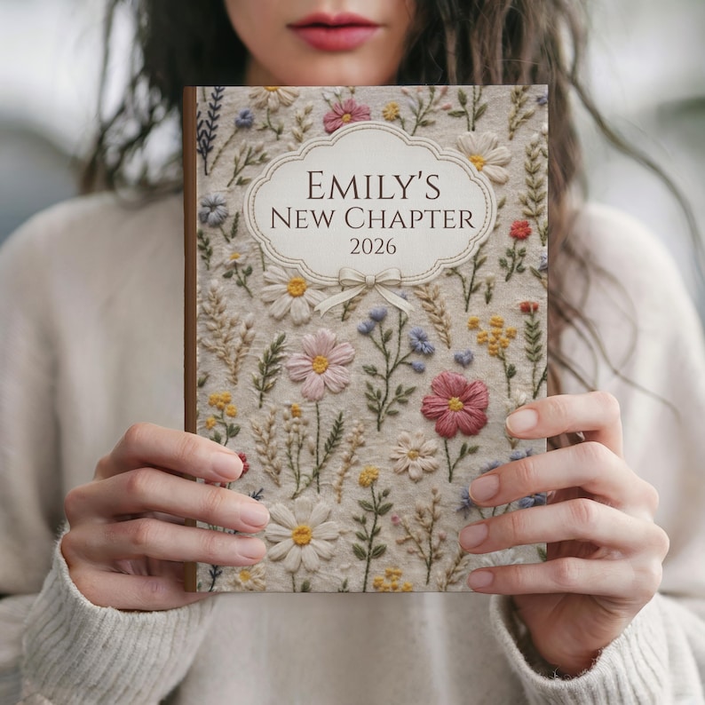 May include: A floral-embroidered journal with the text "Emily's New Chapter 2026" in a decorative frame. The cover features a cream-colored background with colorful embroidered flowers and greenery. The journal has a wooden spine.
