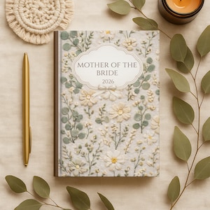 May include: A floral-themed journal with the text "MOTHER OF THE BRIDE 2026" on the cover. The journal features a cream-colored background with embroidered flowers and greenery. A gold pen and a woven coaster are next to the journal.