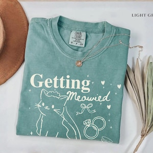 Puede incluir: Camiseta verde claro con la frase "Getting Meowied" y una ilustraci&oacute;n de gato. La camiseta tambi&eacute;n presenta un lazo, corazones y anillos de boda. Un collar dorado est&aacute; drapeado sobre la camiseta.