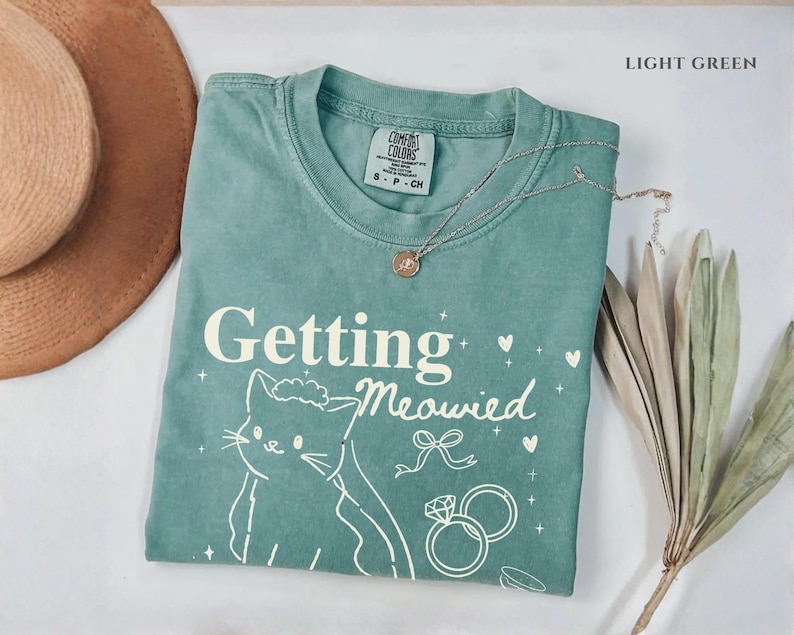 Puede incluir: Camiseta verde claro con la frase "Getting Meowied" y una ilustraci&oacute;n de gato. La camiseta tambi&eacute;n presenta un lazo, corazones y anillos de boda. Un collar dorado est&aacute; drapeado sobre la camiseta.