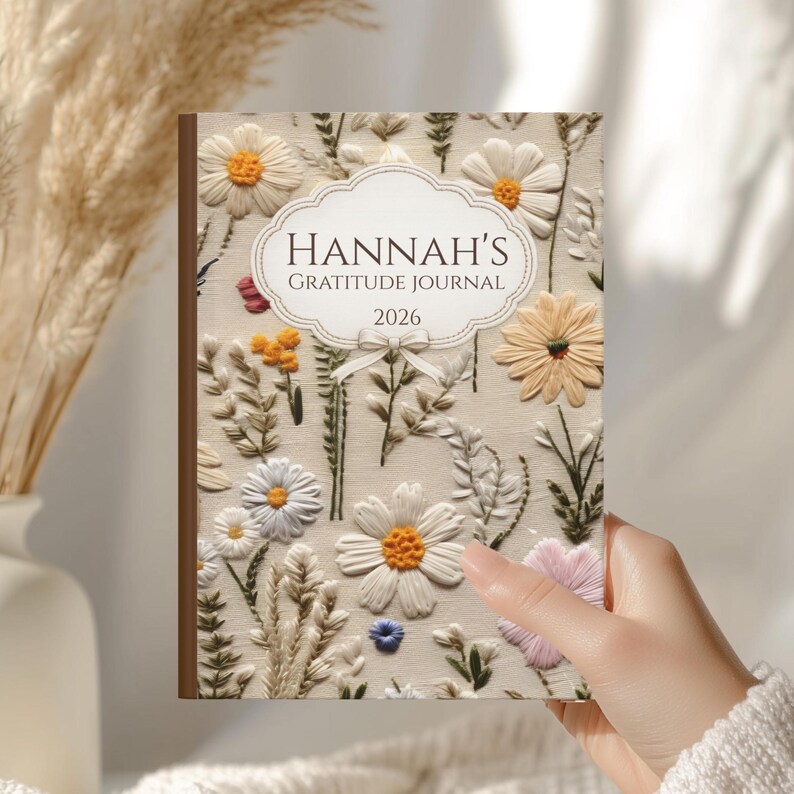May include: A journal with a brown spine and a cover featuring embroidered flowers in white, yellow, and pink. The cover reads "HANNAH'S GRATITUDE JOURNAL 2026" in a decorative font, with a cream-colored background.