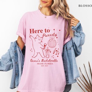 Puede incluir: Camiseta rosa claro con un gr&aacute;fico rojo que dice "Here to Purrrty" con un gato sosteniendo una copa de vino. El texto "Emma's Bachelorette, Miami-Florida, Est. 2025" tambi&eacute;n est&aacute; impreso. La camiseta es de corte holgado.