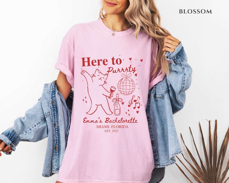 Puede incluir: Camiseta rosa claro con un gr&aacute;fico rojo que dice "Here to Purrrty" con un gato sosteniendo una copa de vino. El texto "Emma's Bachelorette, Miami-Florida, Est. 2025" tambi&eacute;n est&aacute; impreso. La camiseta es de corte holgado.