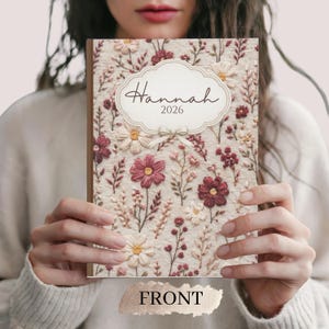 May include: A floral-patterned journal with the name "Hannah" and the year "2026" on the cover. The cover features a cream-colored background with embroidered flowers in shades of burgundy, pink, and white. The word "FRONT" is printed at the bottom.