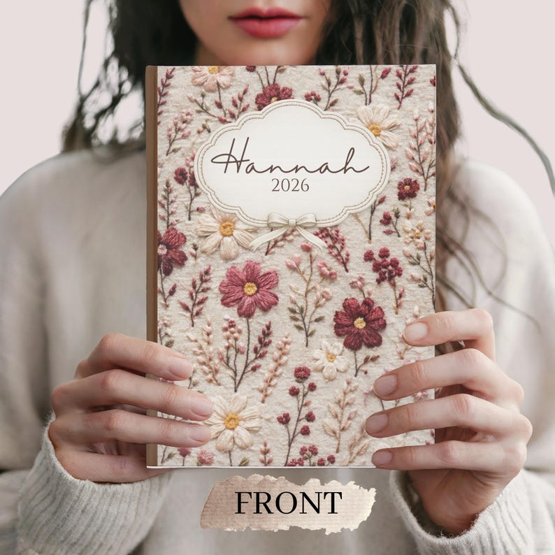 May include: A floral-patterned journal with the name "Hannah" and the year "2026" on the cover. The cover features a cream-colored background with embroidered flowers in shades of burgundy, pink, and white. The word "FRONT" is printed at the bottom.