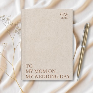 May include: A beige journal with the text "GW 2025" and "TO MY MOM ON MY WEDDING DAY" printed on the front. A gold pen is lying on a white surface next to the journal.