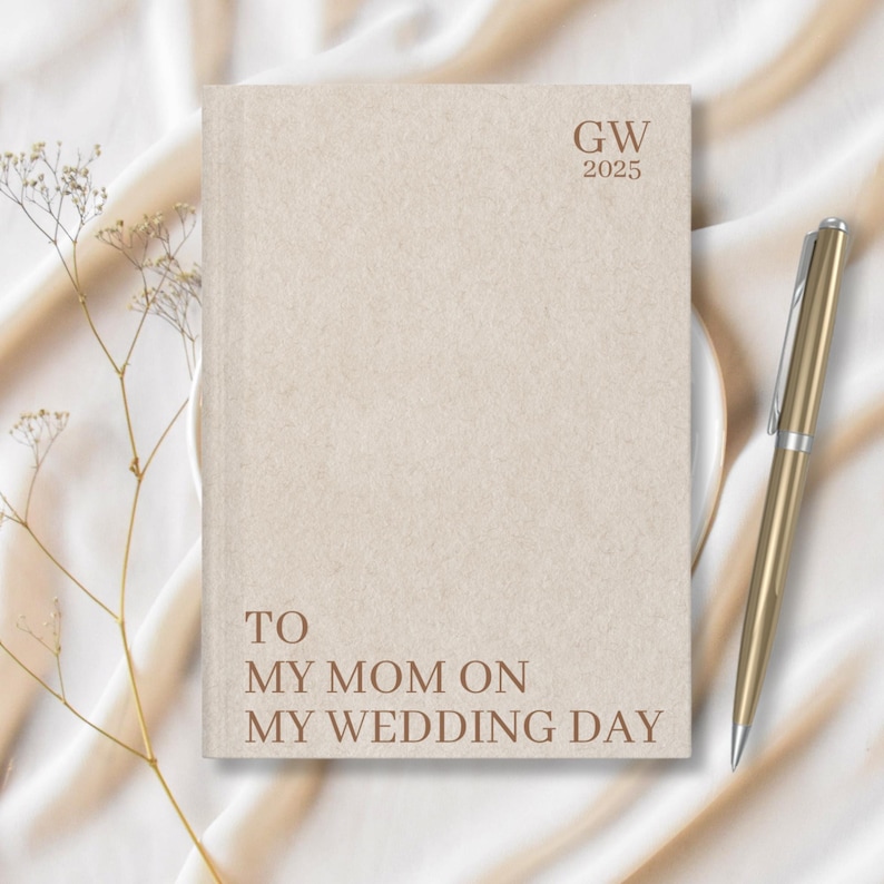 May include: A beige journal with the text "GW 2025" and "TO MY MOM ON MY WEDDING DAY" printed on the front. A gold pen is lying on a white surface next to the journal.