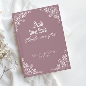 May include: A mauve-colored book with the text "And they lived Happily ever after" in white script. The names "ABIGAIL & FRANK" and the date "03-17-24" are also printed on the cover. White floral corner designs add a decorative touch.