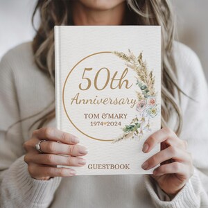 May include: A white guest book with a gold circle and the text "50th Anniversary" with the names "Tom & Mary" and the dates "1974&hearts;2024". The circle is decorated with a watercolor illustration of dried grasses and flowers.