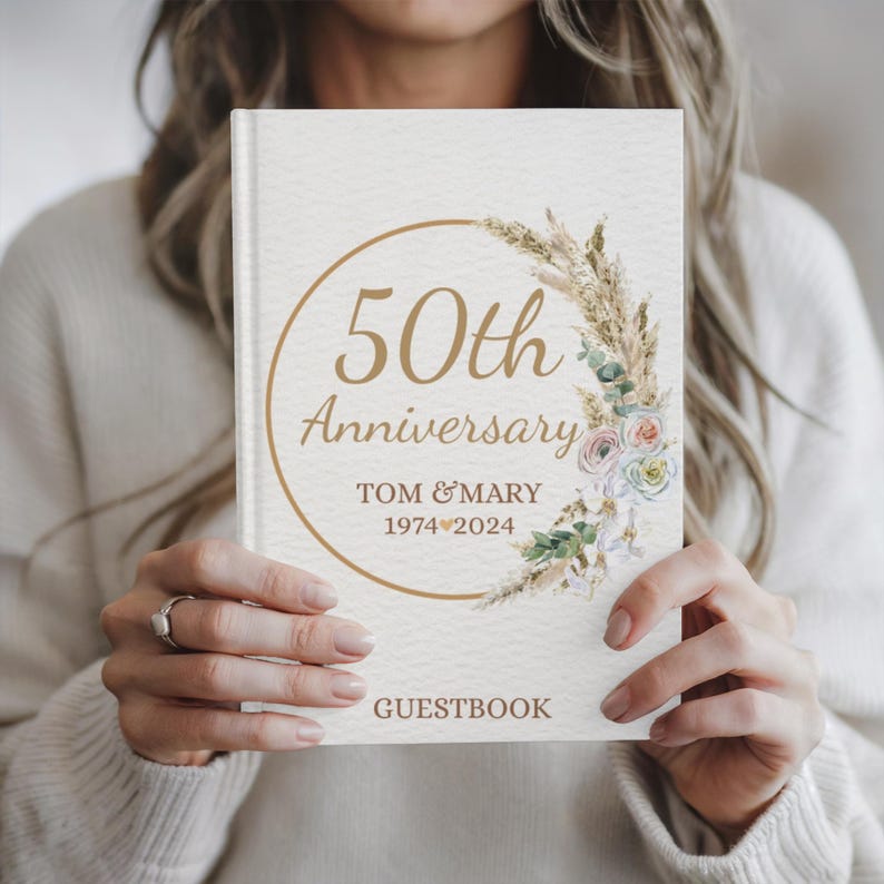 May include: A white guest book with a gold circle and the text "50th Anniversary" with the names "Tom & Mary" and the dates "1974&hearts;2024". The circle is decorated with a watercolor illustration of dried grasses and flowers.