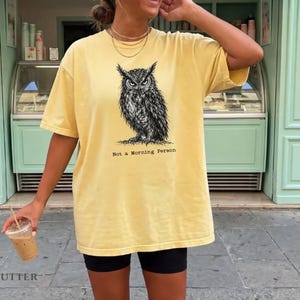 Puede incluir: Una camiseta de manga corta color amarillo mantequilla con una ilustraci&oacute;n detallada en tinta negra de un b&uacute;ho centrada en el pecho. Debajo del b&uacute;ho, el texto Not a Morning Person aparece en una fuente estilo m&aacute;quina de escribir.