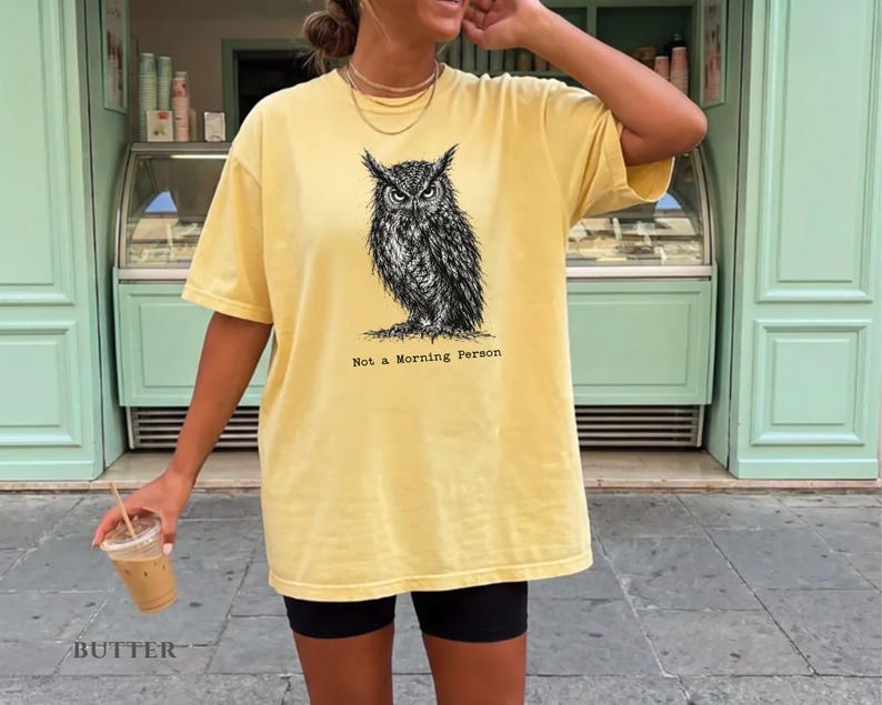 Puede incluir: Una camiseta de manga corta color amarillo mantequilla con una ilustraci&oacute;n detallada en tinta negra de un b&uacute;ho centrada en el pecho. Debajo del b&uacute;ho, el texto Not a Morning Person aparece en una fuente estilo m&aacute;quina de escribir.