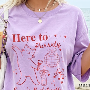 Puede incluir: Camiseta lila claro con el texto rojo "Here to Purrty", una ilustraci&oacute;n de gato sosteniendo una copa de vino, una bola de discoteca, una botella de champ&aacute;n, notas musicales y corazones. La palabra "ORCHID" est&aacute; impresa en la parte inferior derecha.
