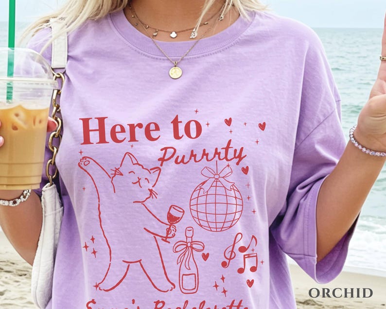 Puede incluir: Camiseta lila claro con el texto rojo "Here to Purrty", una ilustraci&oacute;n de gato sosteniendo una copa de vino, una bola de discoteca, una botella de champ&aacute;n, notas musicales y corazones. La palabra "ORCHID" est&aacute; impresa en la parte inferior derecha.
