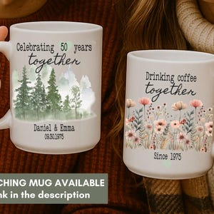 Może przedstawiać: Dwa białe ceramiczne kubki z uchwytami. Jeden kubek m&oacute;wi "Celebrating 50 years together" z leśną scenerią. Drugi m&oacute;wi "Drinking coffee together" z kwiatami. Oba kubki mają daty. Dostępny jest pasujący kubek.