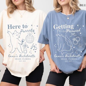 Puede incluir: Dos camisetas, una marfil y otra azul, con dise&ntilde;os de despedida de soltera con tem&aacute;tica de gatos. La camiseta marfil dice "Here to Purrrty" con un gr&aacute;fico de gato, mientras que la camiseta azul dice "Getting Meowied". Ambas camisetas incluyen "Emma's Bachelorette" y la ubicaci&oacute;n y el a&ntilde;o.