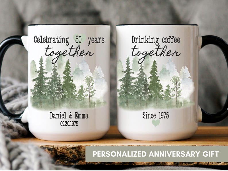 Może przedstawiać: Dwa białe ceramiczne kubki z czarnymi uchwytami. Na jednym kubku napis "Celebrating 50 years together", a na drugim "Drinking coffee together". Oba kubki mają akwarelowy wz&oacute;r lasu. Na spodzie kubk&oacute;w napis "Daniel & Emma 09.30.1975" i "Since 1975".
