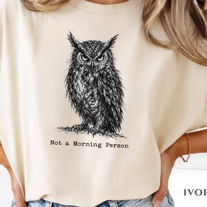 Puede incluir: Una camiseta de color marfil con una ilustraci&oacute;n detallada en tinta negra de un b&uacute;ho con expresi&oacute;n severa. Debajo del b&uacute;ho, el texto "Not a Morning Person" aparece en una fuente estilo m&aacute;quina de escribir. Cuello redondo y mangas cortas.