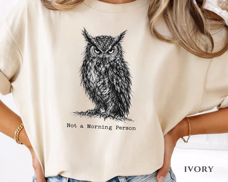 Puede incluir: Una camiseta de color marfil con una ilustraci&oacute;n detallada en tinta negra de un b&uacute;ho con expresi&oacute;n severa. Debajo del b&uacute;ho, el texto "Not a Morning Person" aparece en una fuente estilo m&aacute;quina de escribir. Cuello redondo y mangas cortas.
