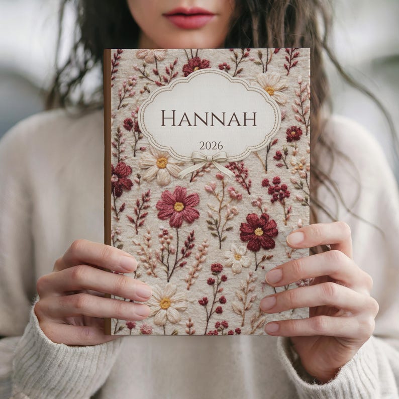 May include: A personalized journal with a floral embroidered cover. The cover features a cream-colored background with embroidered flowers in shades of burgundy, pink, and cream. The name "HANNAH" and the year "2026" are displayed on a decorative label.