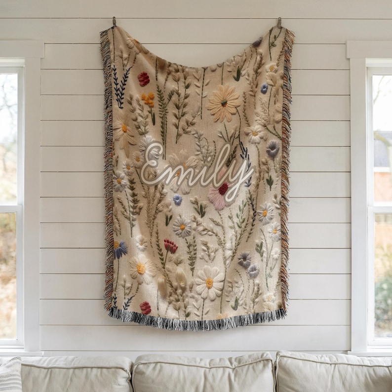 May include: Beige throw blanket with embroidered floral design and the name "Emily" in white script. The blanket features a variety of colorful flowers and foliage, with a fringed edge. The blanket is hanging on a white wall.
