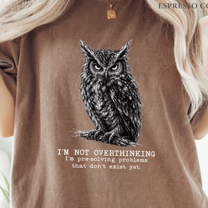 Puede incluir: Una camiseta color espresso con una ilustraci&oacute;n detallada en blanco y negro de un b&uacute;ho posado en una rama. Debajo del b&uacute;ho, el texto dice: I'M NOT OVERTHINKING I'm pre-solving problems that don't exist yet. La camiseta tiene cuello redondo.