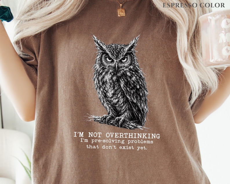Puede incluir: Una camiseta color espresso con una ilustraci&oacute;n detallada en blanco y negro de un b&uacute;ho posado en una rama. Debajo del b&uacute;ho, el texto dice: I'M NOT OVERTHINKING I'm pre-solving problems that don't exist yet. La camiseta tiene cuello redondo.