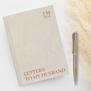 May include: A beige journal with the text "LETTERS TO MY HUSBAND" and the initials "JM" and the year "2024" printed on the front cover. A silver pen is lying next to the journal.