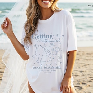 Puede incluir: Camiseta blanca con una ilustraci&oacute;n de gato azul claro y el texto "Getting Meowied". La camiseta tambi&eacute;n incluye el texto "Emma's Bachelorette, Miami-Florida, Est. 2023". Tema de despedida de soltera.