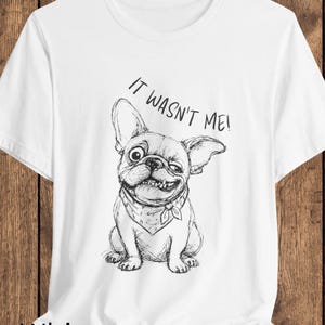 Könnte beinhalten: Ein weißes T-Shirt mit kurzen Ärmeln und einer schwarzen Strichzeichnung einer Französischen Bulldogge mit Halstuch. Der Hund hat einen schelmischen Ausdruck, wobei ein Auge größer ist als das andere. Darüber steht der Text IT WASN'T ME!