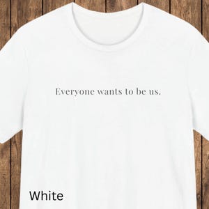 May include: White t-shirt with the text "Everyone wants to be us." printed in black. The word "White" is printed in black at the bottom of the shirt. The shirt has a crew neck and short sleeves.