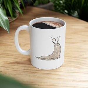 May include: White ceramic coffee mug with a cartoon illustration of a brown slug with large eyes. The mug is filled with coffee.