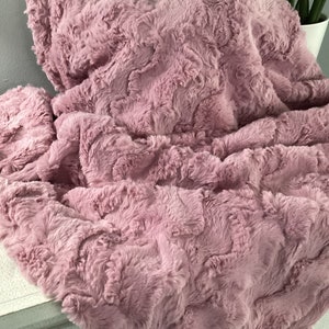 May include: A soft, plush throw blanket in a dusty rose color. The blanket has a textured, fluffy surface and is made of faux fur.