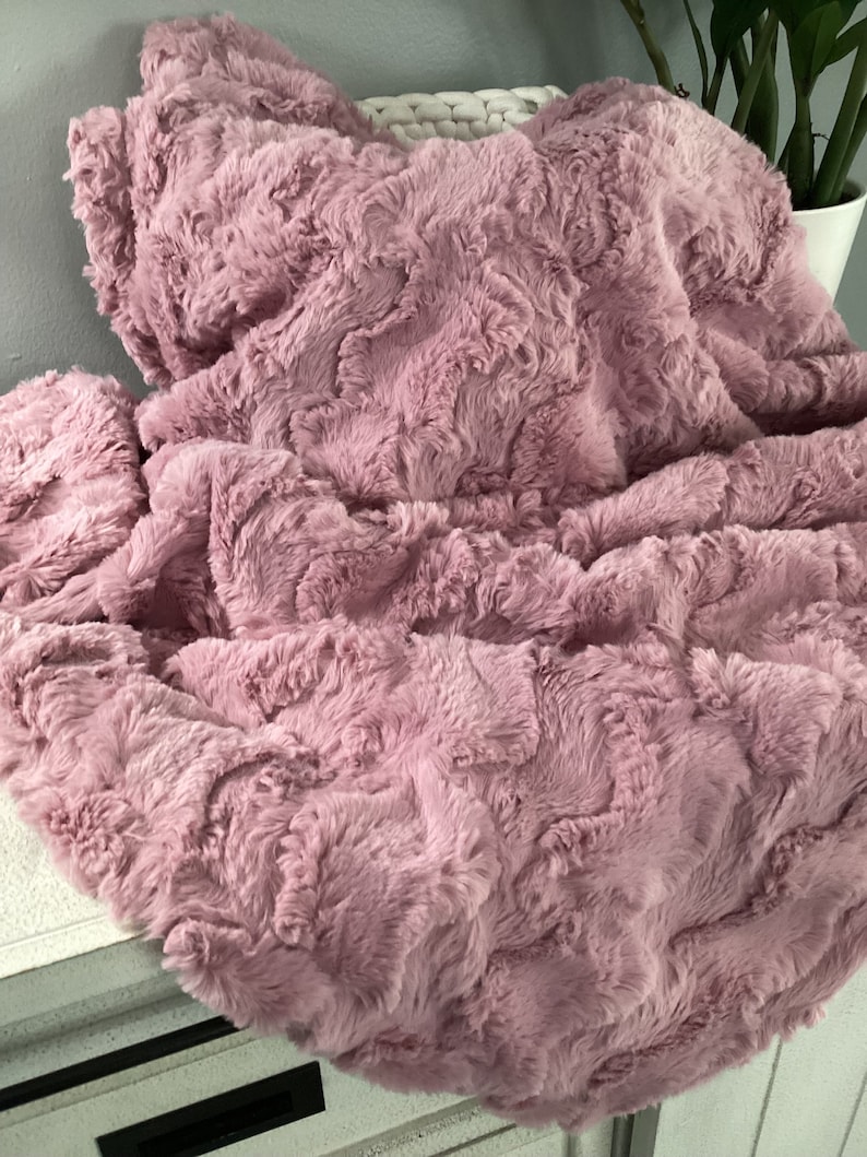 May include: A soft, plush throw blanket in a dusty rose color. The blanket has a textured, fluffy surface and is made of faux fur.