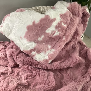 May include: A soft, pink and white faux fur throw blanket with a tie-dye pattern. The blanket is folded and draped over a wicker basket.