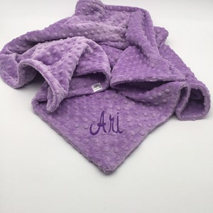 May include: A soft, purple, plush blanket with a textured dot pattern. The blanket is folded and has the name "Ari" embroidered on it.