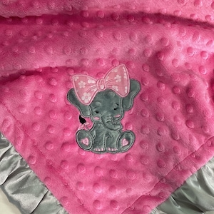 Puede incluir: Manta de bebé rosa con un elefante bordado gris que lleva un lazo rosa. La manta tiene un ribete de satén gris.
