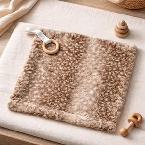 May include: A soft, square baby blanket in shades of brown and tan, with a plush, textured surface. It features a wooden ring attached with a white fabric loop. A small wooden stacking toy and a wooden rattle are also present.