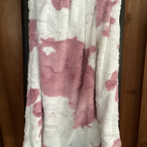 May include: A soft, plush throw blanket with a pink and white cow print design. The blanket is folded and hanging on a wooden rack.