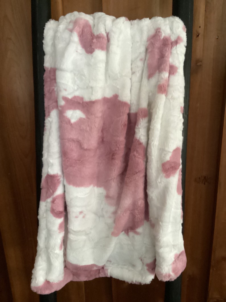 May include: A soft, plush throw blanket with a pink and white cow print design. The blanket is folded and hanging on a wooden rack.